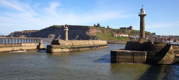 This landscape photograph shows Whitby Harbour in Yorkshire during a clear spring afternoon. The scene prominently features the two lighthouses at the entrance of the harbour, with the historic ruins of Whitby Abbey and St Mary’s Church visible on the hilltop in the background. The sunlight highlights the stone structure of the piers and lighthouses, while the houses of Whitby are clustered along the shoreline. St Mary’s Church stands out amongst the buildings on the green hillside, and Whitby Abbey’s ruins provide a dramatic focal point above the town. The image captures the maritime character of Whitby, showcasing the coastal heritage of Yorkshire with its harbour and lighthouse, and the distinctive architecture of the church and abbey overlooking the waterfront.